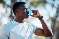 A person in a white shirt uses a blue inhaler outdoors. They stand against a blurred background of trees, with sunlight filtering through. They wear a wristwatch and have a focused expression.