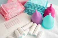 A variety of menstrual products are displayed, including pink and blue wrapped pads, white tampons, and reusable menstrual cups in blue, purple, and pink, all arranged on a soft, white cloth.