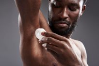A shirtless man applies deodorant to his underarm with a focused expression. The background is a plain gray, highlighting the action. His right hand holds the deodorant stick, and his gaze is directed toward the area of application.