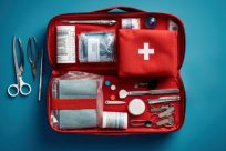 Open red first aid kit on a blue background, containing various medical supplies: scissors, tweezers, bandages, a thermometer, adhesive bandages, gauze, and a red pouch with a white cross. Tools are neatly arranged beside the kit.