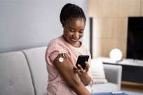 A woman sits on a gray sofa, smiling as she looks at her smartphone. She has a medical sensor patch on her upper arm and wears a pink shirt. A soft lamp and a TV are in the blurred background.