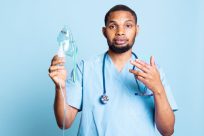 A healthcare professional in blue scrubs holds a nebulizer mask in one hand and gestures with the other. A stethoscope hangs around their neck. The background is a solid light blue.
