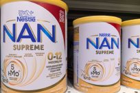 Cans of NAN Supreme infant formula are displayed on a shelf. The labels include details like the age suitability of 0-12 months, immunity benefits, and HMO content. The branding features blue and gold colors with Nestlés logo prominently.